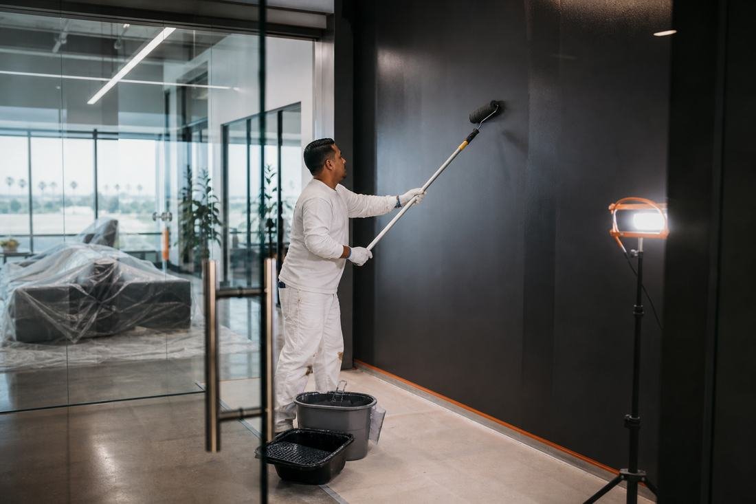A painter rolling a dark accent wall in a glass-walled commercial office, seating sheeted behind him.